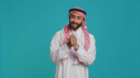 Photo for Hopeful arab man begging and pleading for something, holding hands in a prayer and showing belief. Middle eastern person being desperate and seeking help, having hope in blue studio. - Royalty Free Image