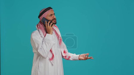 Muslim model answers mobile phone call on wireless device, communicating on remote telephone line. Young person representing islamic tradition with costume and headscarf, answering chat.
