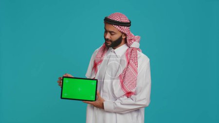 Téléchargez les photos : L'homme musulman présente la tablette d'écran vert sur fond bleu, montrant l'affichage blanc de copyspace. Jeune en vêtements arabes tenant gadget avec modèle de maquette chromakey isolé. - en image libre de droit