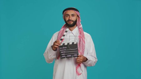 Téléchargez les photos : Le cinéaste moyen-oriental tient un tableau à clins utilisé pour les prises de scène sur la production cinématographique, le réalisateur arabe utilisant l'ardoise de tournage cinématographique. Personne musulmane portant des vêtements arabes traditionnels. - en image libre de droit