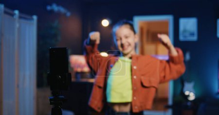 Téléchargez les photos : Enfant talentueux utilisant un smartphone sur un stabilisateur de trépied pour filmer un tutoriel de danse pour d'autres enfants regardant son contenu de médias sociaux en ligne. Joyeux petite fille enregistrement vidéo ciblant la génération Z téléspectateurs - en image libre de droit