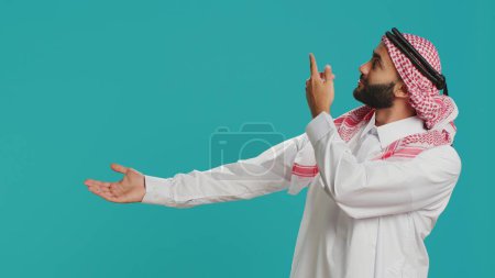 Foto de Hombre musulmán confiado en la ropa tradicional que muestra con las manos en el copyspace para el anuncio comercial en el estudio. Sonriente persona islámica mostrando a un lado con los brazos y mirando a la cámara. - Imagen libre de derechos