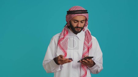 Araber, die Kartennummern auf Webseiten hinzufügen, Online-Shopping auf Shopping-App im Einkaufszentrum machen. Person, die auf Internetseiten bezahlt und traditionell islamische Kleidung mit Schal trägt.