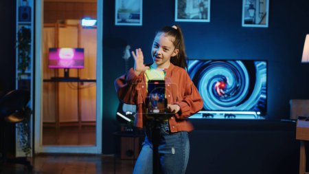Chica joven bailando en el interior del estudio hogar débilmente iluminado, produciendo contenido con el teléfono móvil. Niño sonriente haciendo coreografía de danza viral en la sala de estar con animaciones 3D en monitores de fondo