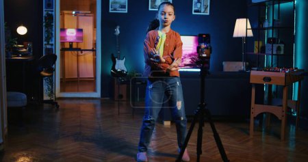 Photo for Panning shot of viral media star dancing in apartment, recording video with phone on tripod for gen z internet fans. Excited little child filming herself with cellphone doing dance moves - Royalty Free Image