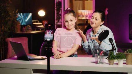 Photo for Young girl and her mother filming content for their internet channel, talking about their relationship. Cute kid and mom recording family vlog in apartment studio with pink neon lighting as background - Royalty Free Image