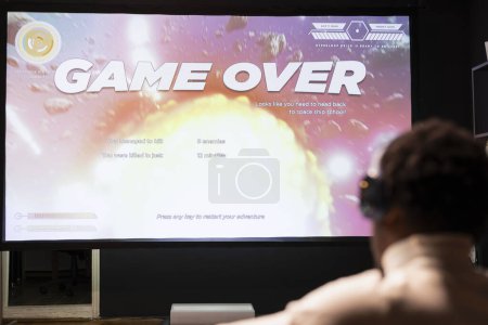 Photo for Gamer wearing headphones upset at seeing game over message on widescreen TV while playing arcade space shooter videogame. Man relaxing at home on gaming system, disappointed after losing - Royalty Free Image