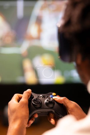 Close up shot of controller held by gamer sitting in front of gigantic television set displaying console game. Joystick used by homeowner enjoying gaming session on smart TV, relaxing in living room