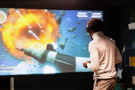 Gamer at home gaming and enjoying leisure time using high quality projector to display gameplay. Man standing up in front of ultrawide screen, playing videogame using controller