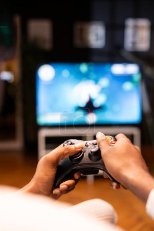 Focus shot on man holding controller in apartment, playing videogames on smart TV display in blurry background. Gamer on sofa using joypad to participate in game on console attached to television set