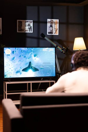 Gamer in front of widescreen smart TV watching SF videogame gameplay. African american man wearing headphones in home theatre to enjoy gaming content online displayed on television set