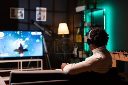 Photo for Man plays science fiction singleplayer game on gaming console while talking with friends through headphones. Player enjoying videogame with high quality graphics, using gamepad to navigate spacecraft - Royalty Free Image