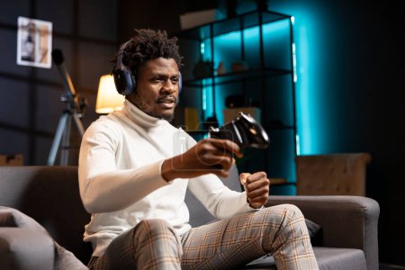 Photo for Man in cyan neon lit apartment playing videogames on gaming console with teammates. BIPOC player fights foes in esports game using joystick, discussing with friends through headset - Royalty Free Image