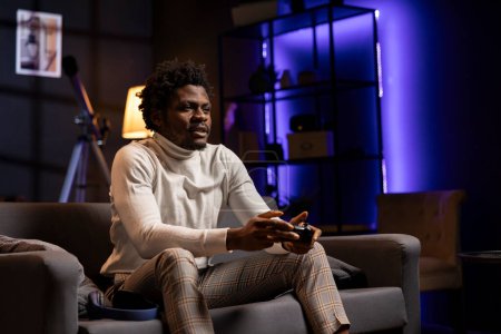 Gamer zu Hause spielen und die Freizeit genießen. Afrikanischer Mann sitzt auf Couch in blauem neonbeleuchtetem Wohnzimmer, spielt Videospiel auf Konsole, entspannt sich nach einem harten Arbeitstag