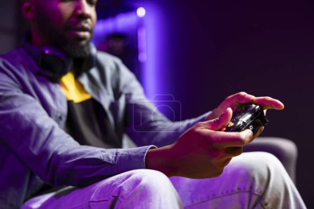 Gamer auf der Couch mit Controller und Kopfhörer, um zu Hause Videospiele aus nächster Nähe zu spielen. Afroamerikaner im Heimkino mit High-Tech-Gamepad der Spielkonsole, um Gegner im Spiel zu besiegen