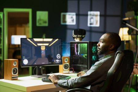 Foto de Hombre afroamericano jugando videojuego FPS en la pantalla del PC en la sala de estar apartamento iluminado de neón. Gamer disfrutando del juego de disparos en la pantalla de escritorio de la computadora en casa iluminada RGB tarde en la noche - Imagen libre de derechos