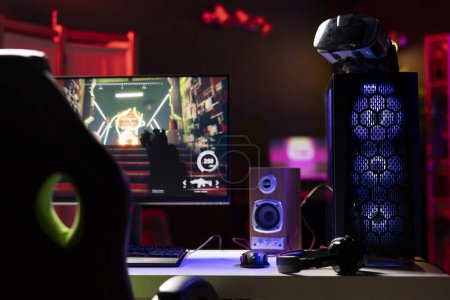 Téléchargez les photos : Studio de salle vide de jeu professionnel avec néons et ordinateur RVB allumé à côté des écouteurs et casque VR. Jeu vidéo FPS sur écran PC et lunettes de réalité virtuelle dans l'appartement - en image libre de droit