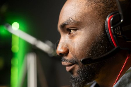 Smiling BIPOC man has fun playing videogames on PC while chatting with teammates through headphones mic. Close up of upbeat african american gamer communicating with friends through headset microphone