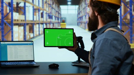 Packaging engineer looking at tablet with greenscreen in depot, working in warehouse with blank copyspace template. Professional depot employee examines device with isolated display.