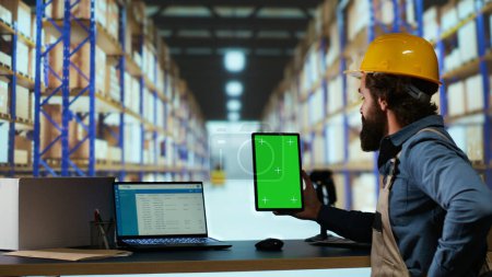 Warehouse administrator has greenscreen on tablet before creating invoices on merchandise parcel distribution, examining isolated chromakey on gadget. Industrial worker checks blank copyspace.