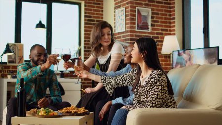 Photo for Multiracial group of friends talking with each other in brick wall apartment living room, gathered together to celebrate holiday. Colleagues socializing at home, enjoying wine and snacks - Royalty Free Image