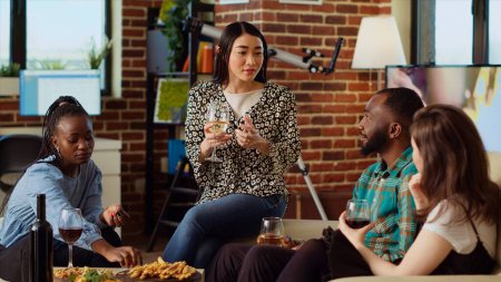 Asian woman telling entertaining story to friends at apartment party during weekend gathering while everyone enjoys wine glass. Multiethnic guests listening to funny tale from host in cozy living room