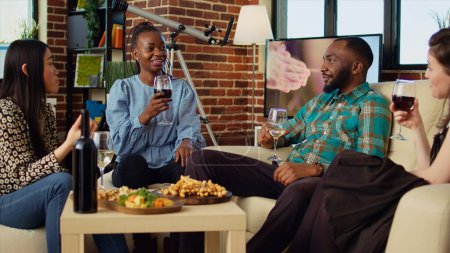 Afroamerikanerin bezaubert Freundinnen auf Wohnungsfeier mit fesselnder Geschichte. Gast bringt multiethnische Gruppe im stilvollen Wohnzimmer zum Lachen, während er trinkt und isst