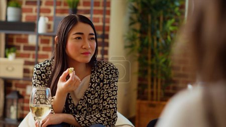 Asian woman paying attention to interesting discussion between multiethnic apartment party guests. Host eating gourmet cheese from charcuterie board and listening to friends conversation