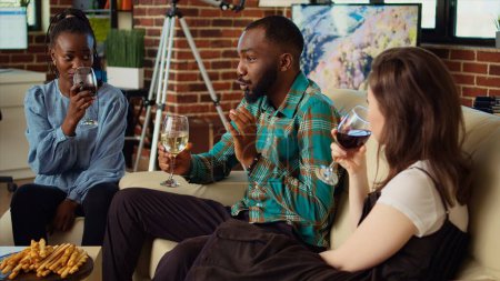 Foto de Diversos amigos del grupo celebran el cumpleaños del hombre afroamericano, beben alcohol y comen aperitivos en el apartamento. Los invitados se ríen y discuten en la reunión de apartamentos después de regalar regalos - Imagen libre de derechos