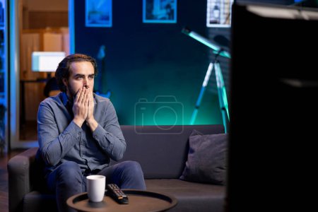 Man watching thriller slasher movie broadcasted on TV in disbelief, shocked by macabre character killing. Person covering mouth with hands, seeing nail biting gruesome film
