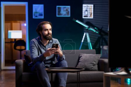 Photo for Man watching TV in apartment and answering business related messages on mobile phone. Teleworking worker on couch in living room looking at movies and responding to texts on smartphone - Royalty Free Image
