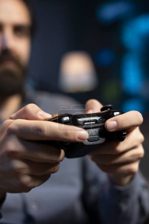 Close up shot of controller held by gamer at home gaming and enjoying leisure time. Focus on gamepad used by man in blurry background in neon lit living room playing videogame on console, relaxing