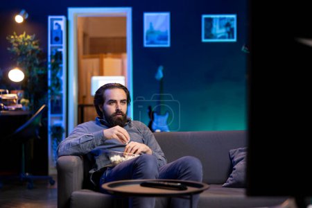 Person using TV to binge series on subscription based streaming services, enjoying bowl of popcorn. Man relaxing in apartment watching VOD shows on television display, eating snack