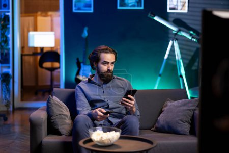 Person in home theater binging series on streaming service, texting friend on phone. Man in neon lit apartment with bowl of popcorn in front watching shows on television and chatting with mate