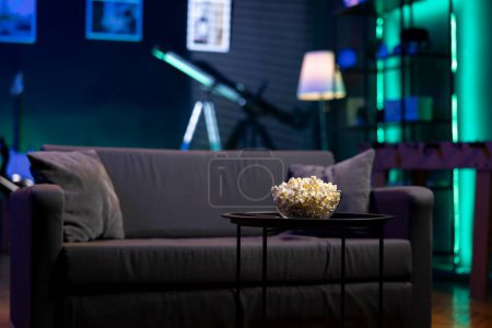 Photo for Bowl of popcorn on coffee table in front of cozy furniture in empty neon lit modern apartment. Snack prepared for movie night in dimly illuminated home theatre in front of couch - Royalty Free Image