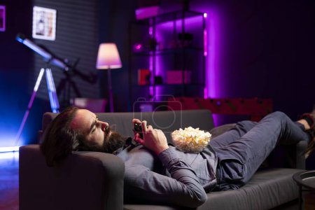 Téléchargez les photos : Gamer s'est allongé sur canapé tout en s'engageant dans un tournoi multijoueur compétitif en ligne en utilisant la console de jeu et la manette de jeu. Homme dans le cinéma maison jouer à des jeux vidéo avec bol de pop-corn dans les genoux - en image libre de droit