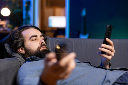 Bored person lounged on couch binging series on streaming services, browsing using remote and texting on phone. Man watching VOD shows on television, zapping though channels, chatting on smartphone