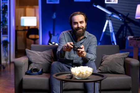 Portrait of smiling man playing videogame and eating snacks, holding controller. Cheerful gamer participating in esports tournament using professional gamepad for better victory chances