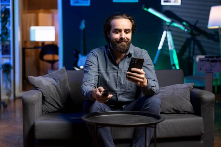 Smiling man watching shows on TV, using remote control, texting friends on smartphone. Cheerful person in pajamas consuming mindless entertainment and chatting with mates on phone