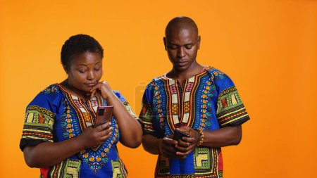 Photo for Ethnic people in traditional clothes checking smartphone apps, looking at messages received on social media network. African american man and woman browsing websites on mobile device. - Royalty Free Image
