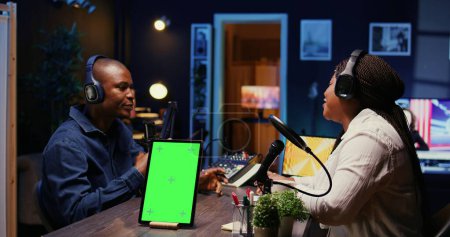 Isolated screen tablet next to vlogger show host chatting with guest in neon lights ornate living room studio. Chroma key device on table with man and woman sitting and discussing on podcast