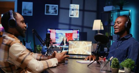 Afroamerikanische Influencer-Aufnahmepodcast mit Gast, mit Audio-Aufnahmegeräten im Heimstudio. Fröhliche Männer plaudern live während der Übertragung der Internet-Show