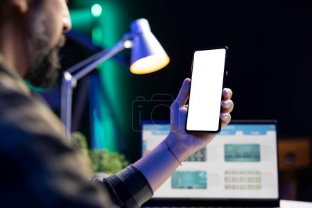 Man vertically holding mobile device with isolated copyspace mockup template. In front of a laptop screen, a male individual is grasping a smartphone showing blank white screen.