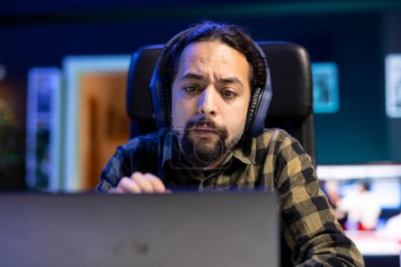 Close up of serious man wearing headphones and looking at his laptop. Male individual working from home, using wireless headset for music while browsing the internet on device.