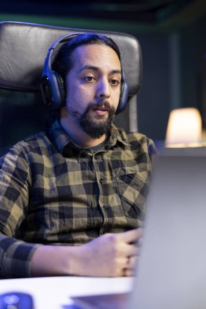 Male individual wearing wireless headphones and looking at his digital laptop. Sitting at a desk is young man utilizing his headset and portable computer for entertainment, watching a movie at home.