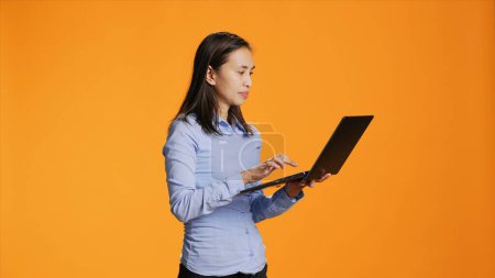 Casual person navigating online on laptop in studio, looking for inspiration on wireless pc against orange background. Asian woman browsing webpages on internet, search project.
