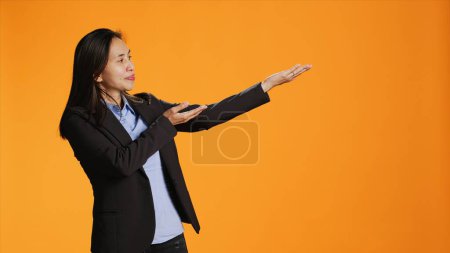 Photo for Asian office worker points to both sides of the background, creating new web promotional ad in orange studio. Young businesswoman working on marketing advertisement during shoot. - Royalty Free Image