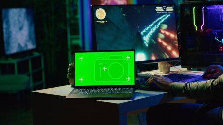 Green-Screen-Laptop neben dem Mann, der Spaß hat, indem er die Gaming-Tastatur benutzt, um im SF-Videospiel Raumschiff zu fliegen. Notebook-Attrappen und Gamer mit leistungsstarken PCs und Computer-Peripherie zum Spielen