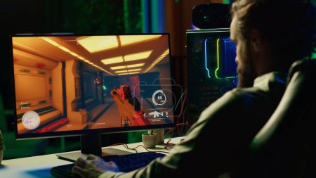 Man in dark living room playing engaging video games on gaming PC at computer desk, chilling after work. Gamer contending against foes in online multiplayer shooter, shooting them with lasers