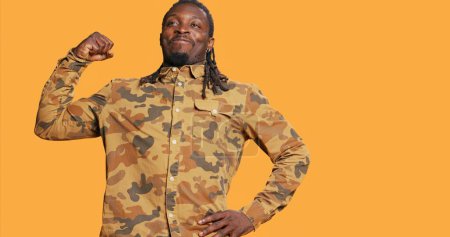 Photo for Athletic strong man flexing his biceps muscles on camera, advertising fitness routine and wellbeing. African american person flex arms and acting powerful over orange background. Handheld shot. - Royalty Free Image